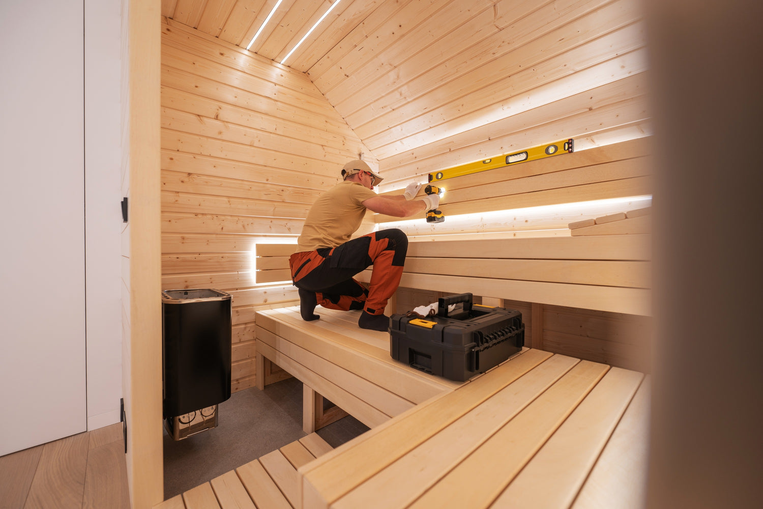 Une personne vêtue d'une chemise beige et d'un pantalon orange travaille à l'intérieur d'un sauna en bois. Elle utilise un outil de niveau pour percer le dossier. Le sauna est composé de bancs et de murs en bois, avec une boîte à outils posée sur l'un des bancs. Un poêle est placé dans un coin.